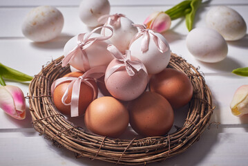 White and brown eggs with pink bows on a nest on a white wooden background and tulip flowers.