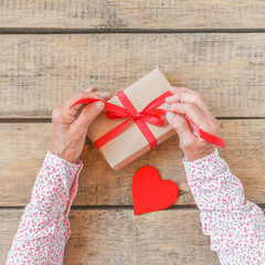 Senior woman packs gift for Valentine day, top view. Wrinkled hands decorate surprise box with red ribbon and heart. Christmas, Thanksgiving, Easter, vintage wooden table. Sharing family tradition.