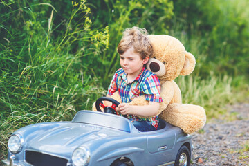 Little preschool kid boy driving big toy car and having fun with playing with his plush toy bear, outdoors. Child enjoying warm summer day in nature landscape.
