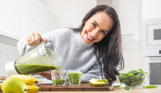 Similing female pours freshly made green smoothie into glasses in the kitchen