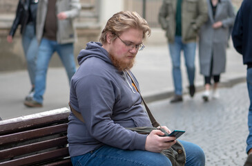 Fototapeta premium Street portrait of a fat young man with glasses and a beard 25-30 years old with blond hair, looking at a mobile phone against the background of old European city houses, people pass by,