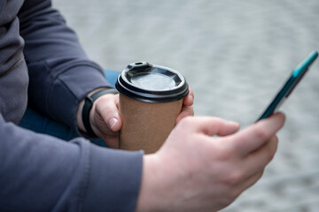 The man looks at his mobile phone and holds a coffee in a paper cup in his hand.