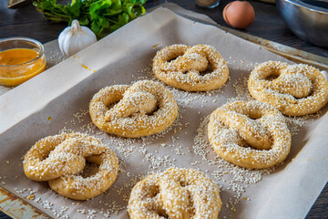 Homemade unbaked soft pretzels with sesame seeds ready to go to the oven in a baking tray. Gluten free dough for savory delicious pastry with a knot. Egg wash, garlic, and coriander in the background