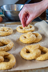 Woman's hand sprinkles sesame seeds on top of homemade unbaked soft pretzels that are ready to go to the oven on a baking sheet. Gluten free dough for savory delicious pastry with a knot. Side view.