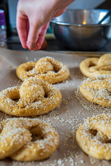 Homemade unbaked soft pretzels ready to go to the oven on a baking sheet. Woman's hand sprinkle sesame on to of pretzels in the background. Gluten free dough for savory delicious pastry with a knot