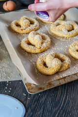 Woman's hand sprinkles garlic flakes on top of homemade unbaked soft pretzels that are ready to go to the oven on a baking sheet. Gluten free dough for savory delicious pastry with a knot. Side view.