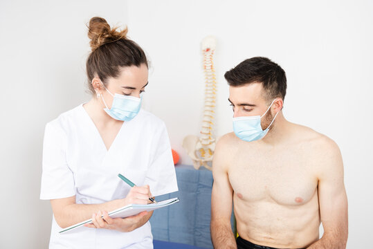 Chiropractor Working With A Patient And Writting On A Notebook While Wearing Face Masks In A Physiotherapy Center During Coronavirus Pandemic.