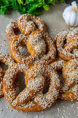 Freshly baked homemade soft pretzel with sesame seeds and garlic flakes, ready to eat. Tasty gluten free savory pastry with a knot on a baking sheet. Coriander and garlic knob in background. Side view