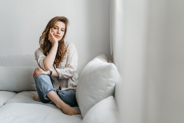 Нoung woman is sitting on the sofa in the living room. A beautiful blonde in white comfortable home clothes.