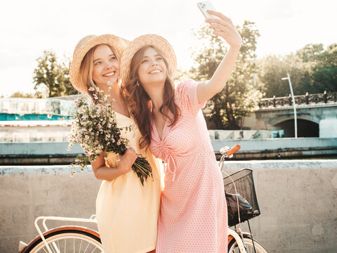 Two young beautiful smiling hipster woman in trendy summer sundress. Sexy carefree women posing on the street background in hats. Positive models at embankment at sunset near bicycle.Taking selfie