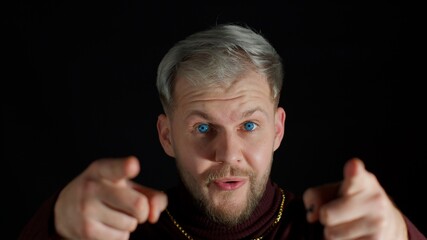 Glad happy stylish young man with blue eyes in trendy clothes smiling excitedly and pointing to camera, beauty choosing lucky winner, indicating to awesome you on black studio background. Hey you