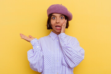 Young mixed race woman wearing a beret isolated on yellow background holds copy space on a palm, keep hand over cheek. Amazed and delighted.