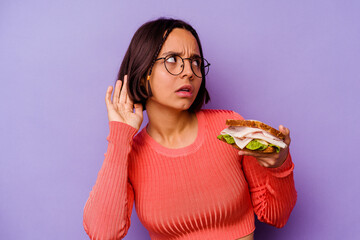 Young mixed race woman holding a sandwich isolated on purple background trying to listening a gossip.