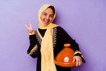 Young Moroccan woman holding a tajine isolated on purple background joyful and carefree showing a peace symbol with fingers.