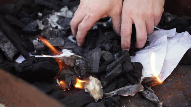 A young man lights a fire with paper matches and coals. Metal grill. Rest in nature, patio Bonfire for cooking barbecue dangerously