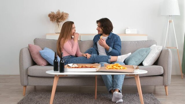 Young happy caucasian couple enjoying time together at home, eating chips with beer and pizza, resting on sofa at home