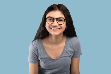 Cute Indian teenage girl in casual outfit and glasses looking at camera on blue studio background