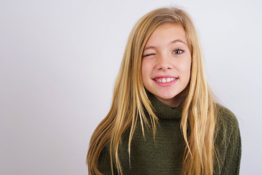 Coquettish Caucasian kid girl wearing green knitted sweater against white wall smiling happily, blinking at camera in a playful manner, flirting with you.