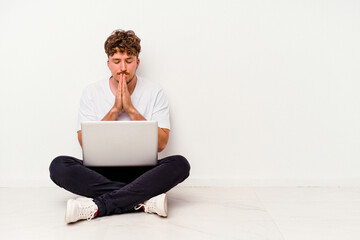 Young caucasian man sitting on the floor holding on laptop isolated on white background holding hands in pray near mouth, feels confident.