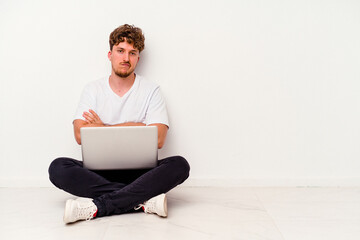 Young caucasian man sitting on the floor holding on laptop isolated on white background unhappy looking in camera with sarcastic expression.