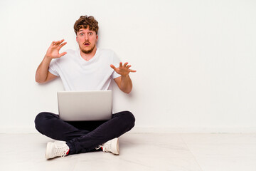 Young caucasian man sitting on the floor holding on laptop isolated on white background being shocked due to an imminent danger