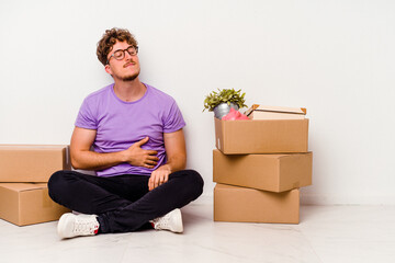 Young caucasian man sitting on the floor ready for moving isolated on white background touches tummy, smiles gently, eating and satisfaction concept.