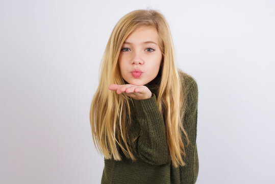 Caucasian kid girl wearing green knitted sweater against white wall looking at the camera blowing a kiss with hand on air being lovely and sexy. Love expression.