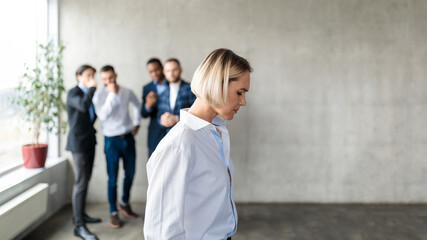 Male Colleagues Whispering Behind Back Of Unhappy Business Lady Indoors - obrazy, fototapety, plakaty
