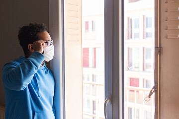 Fototapeta premium Coronavirus Sick man from corona virus looking out the window and wearing mask for protection and recovery from illness at home. Quarantine. Isolated patient to prevent infection. Pandemic. House.