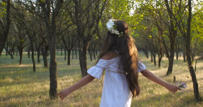 Tracking shot of a  Caucasian teenager girl with long brown hair and blue eyes in a flower wreath dancing beautiful dance of flowers on a nature background. 4k 50 fps slow motion