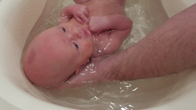 Father holding baby in bath bathing and washing, 1 month old infant in plastic tub in bathroom at home. High quality 4k footage