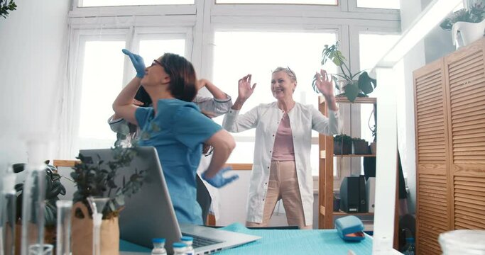 Happy fun excited multiethnic female doctors dance together celebrating victory over coronavirus at clinic workplace.