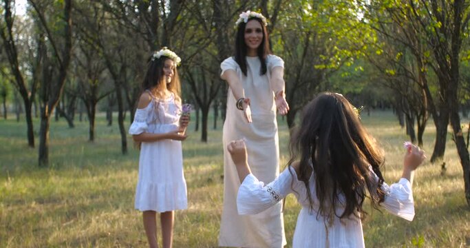 Tracking shot  mother walking with her daughters wearing flower wreaths and white boho dresses  on the grass on a nature background. Mother’s Day concept. 4k 50 fps slow motion