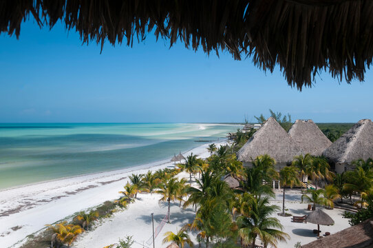 Panoramic view of a beach on Holbox Island in Mexico, with white sand, palm trees, and thatched huts. The perfect destination