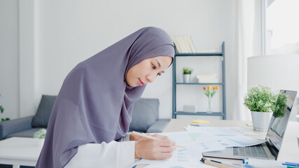 Beautiful Asia muslim lady in headscarf casual wear using laptop in living room at house. Remotely working from home, new normal lifestyle, social distancing, quarantine for corona virus prevention.