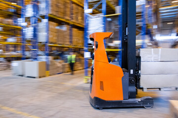 Warehouse worker driving a forklift truck full of goods between high blue yellow steel shelves...