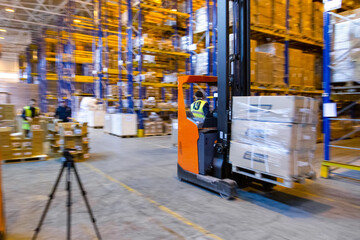Warehouse worker driving a forklift truck full of goods between high blue yellow steel shelves inside warehouse © Roman