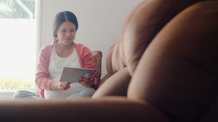 Obraz premium Young Asian Pregnant woman using tablet search pregnancy information. Mom feeling happy smiling positive and peaceful while take care her child lying on sofa in living room at home concept.