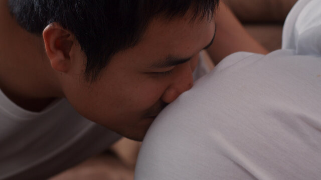 Young Asian Pregnant Couple Man Kissing His Wife Belly Talking With His Child. Mom And Dad Feeling Happy Smiling Peaceful While Take Care Baby, Pregnancy Lying On Sofa In Living Room At Home Concept.