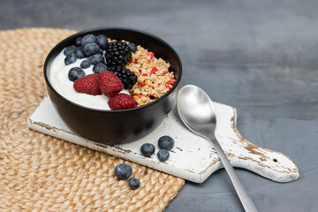Muesli with yougurt and fresh berries in black bowl on dark gray background.
