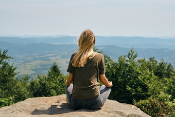 Naklejka na ściany i meble A woman is practicing meditation on a rock with a beautiful view of the mountains. Outdoor adventure. Healthy lifestyle, active rest. Tourism, travel and climbing concept. Enjoying the nature