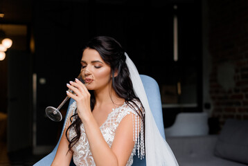 Close up of young brunette bride sitting on a cross and drinking champagne in a hotel room. Concept of bride morning