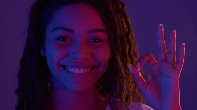 Close up portrait of cute african american woman showing OK gesture and smiling at camera, neon lights background