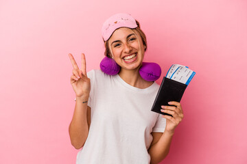 Young caucasian blonde woman holding a passport and tickets to travel isolated on pink background joyful and carefree showing a peace symbol with fingers.