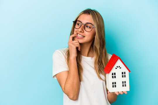 Young caucasian woman holding a house model isolated on blue background relaxed thinking about something looking at a copy space.
