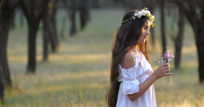 Portrait of a  Caucasian teenager girl with long brown hair and blue eyes in a flower wreath dancing and smelling bouquet of flowers on a nature background. 4k 50 fps slow motion