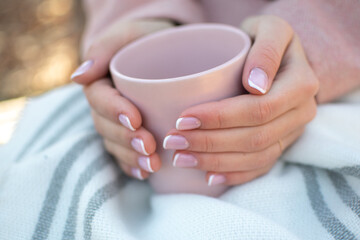 The girl warms her hands with a cup of coffee. Autumn theme. A woman in a pink coat with a beautiful French manicure holds a pink cup in nature.