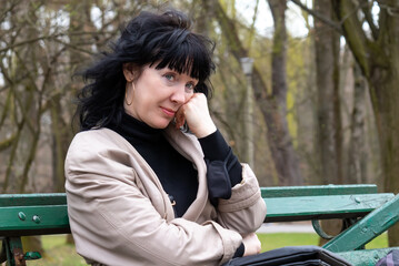 female portrait of a beautiful middle-aged woman with long dark hair sits in a spring park on a bench, resting and with a pensive look