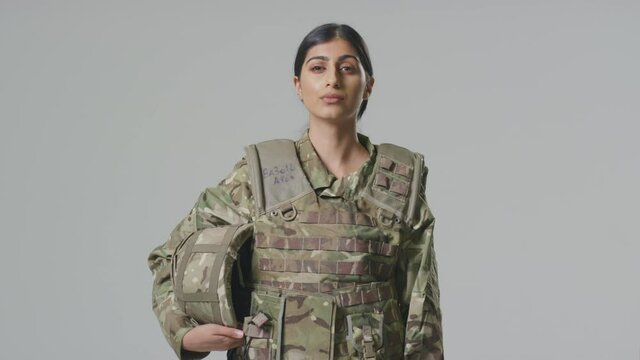 Young female soldier in uniform wearing body armour and holding helmet in front of plain studio background - shot in slow motion