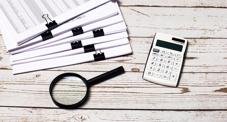 On a light wooden table, there are documents, a magnifying glass, stacks of documents, and a white...